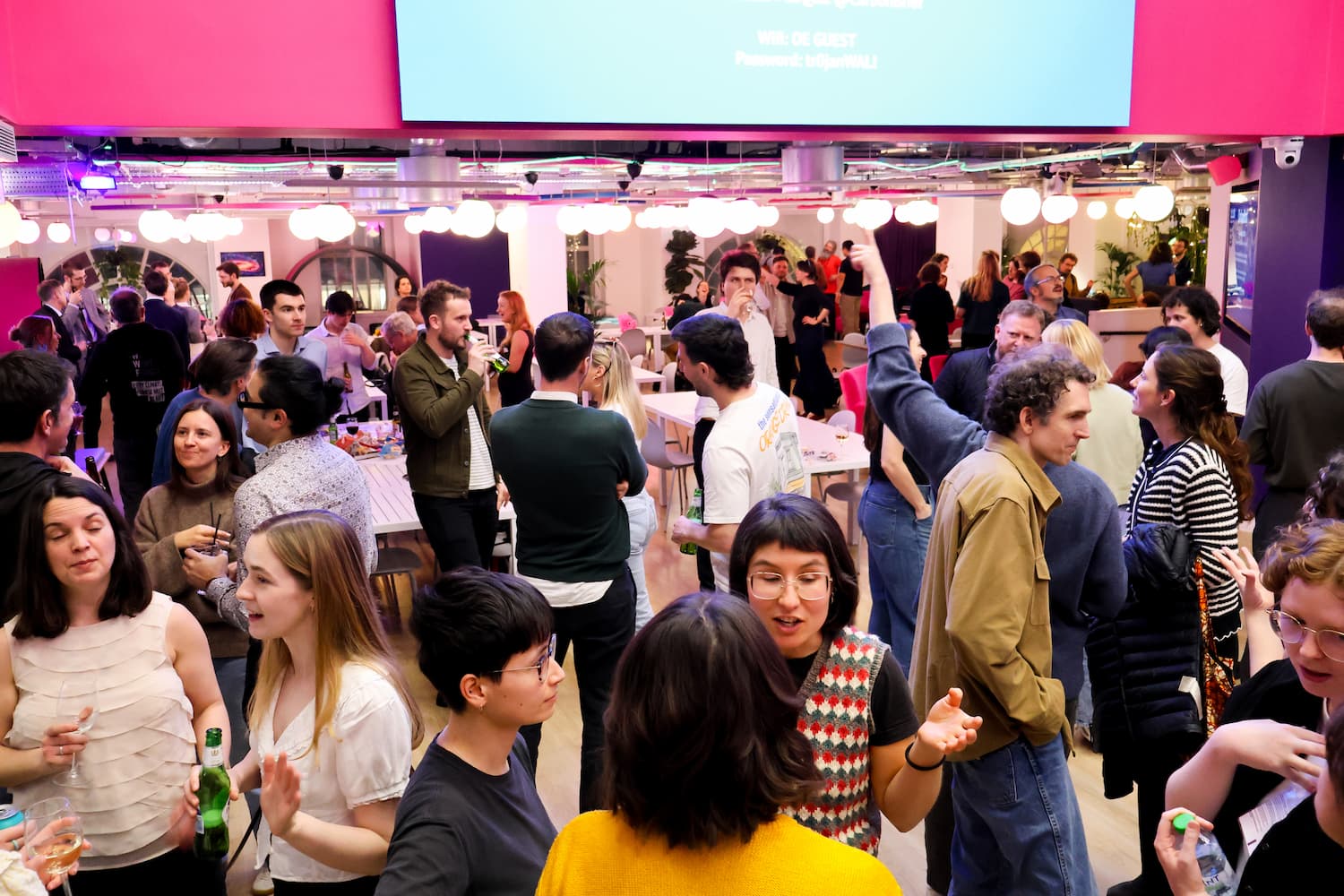 A large, crowded office social space with pink accents. Dozens of people are standing and talking, many holding drinks. A large screen displays Wi-Fi information. The room is brightly lit with round pendant lights.