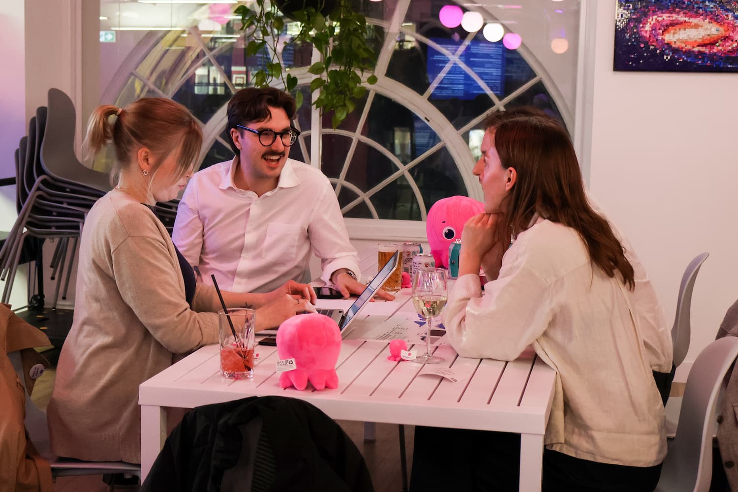 Three people sit around a white table in an office setting, focused on a laptop. A large circular window is in the background. Two small, bright pink plush octopuses sit on the table among drinks.