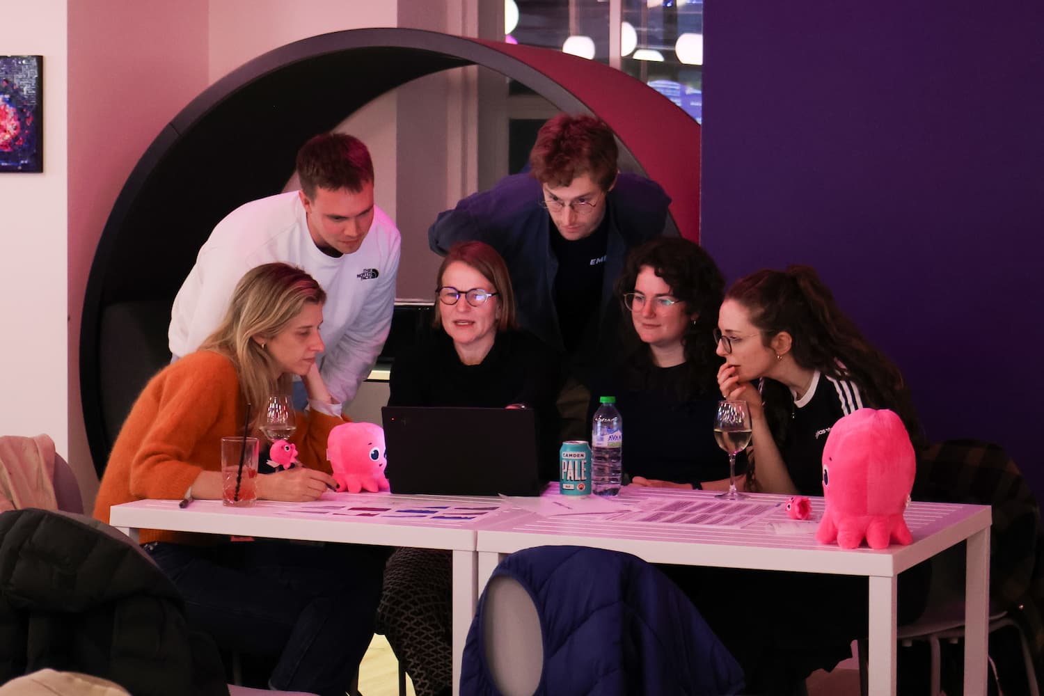 Five people are huddled closely around a laptop on a white table, looking intently at the screen. Several pink octopus plushes and drink cans are scattered on the table. A purple wall is visible to the right.