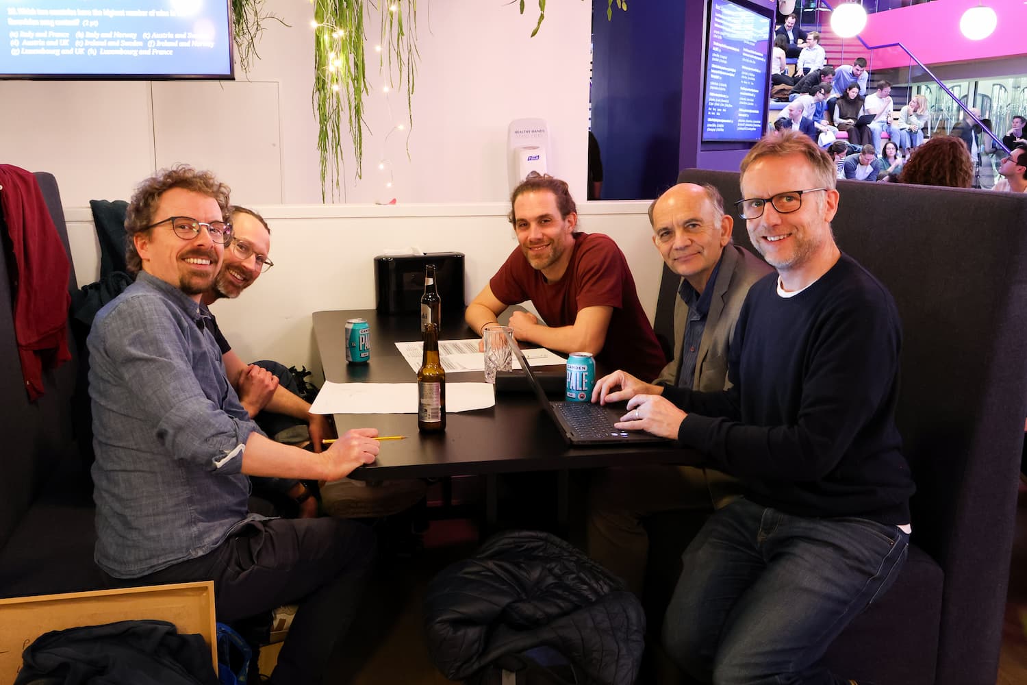 Five men sit in a dark grey booth, smiling toward the camera. They have a laptop, papers, and beer bottles on the table. A screen in the background shows a Eurovision trivia question.
