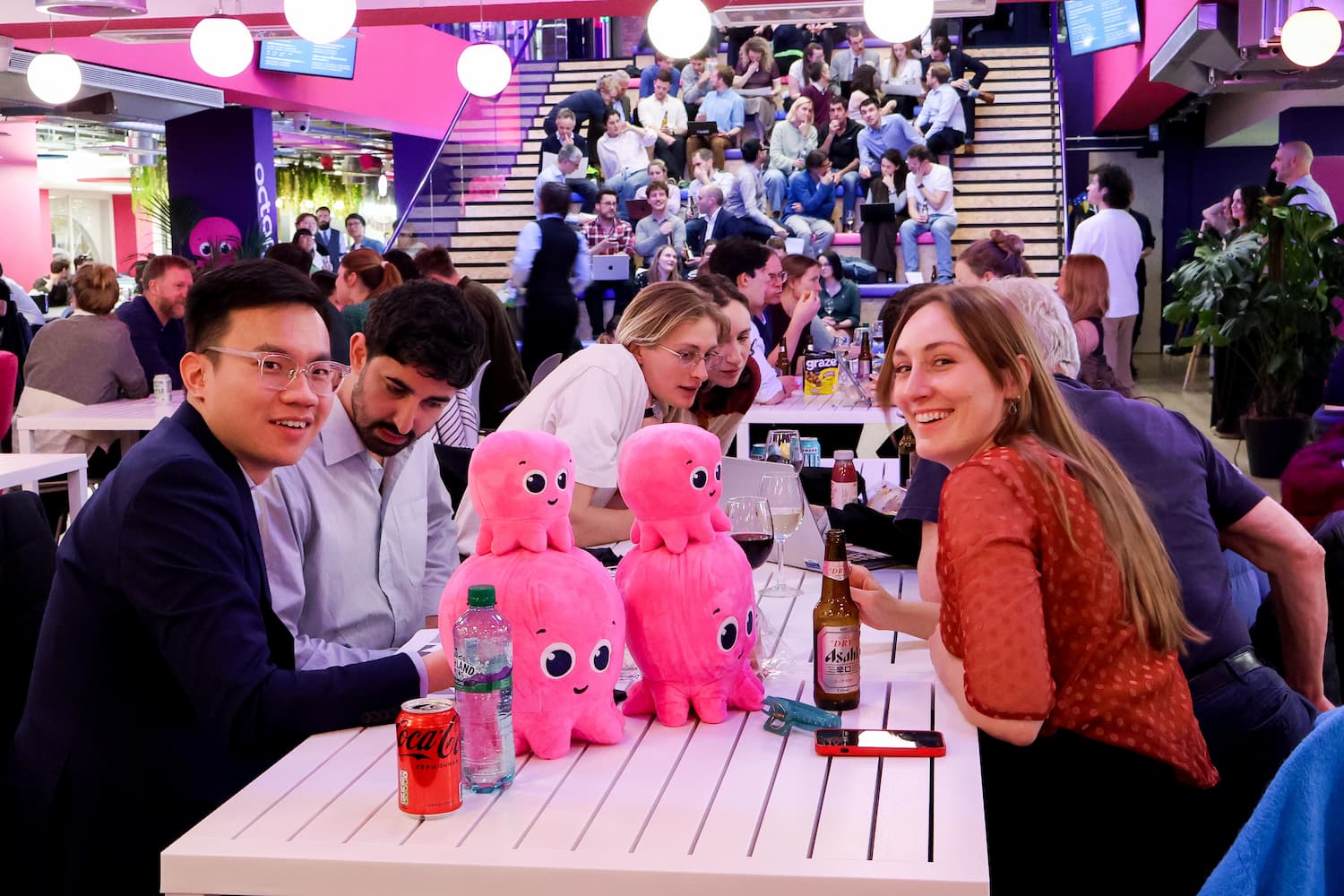 A group of people sits at a long white table. In the foreground, a man in a blue blazer and a woman in a red top smile at the camera. Two large pink octopus plushes are stacked in the center of the table.