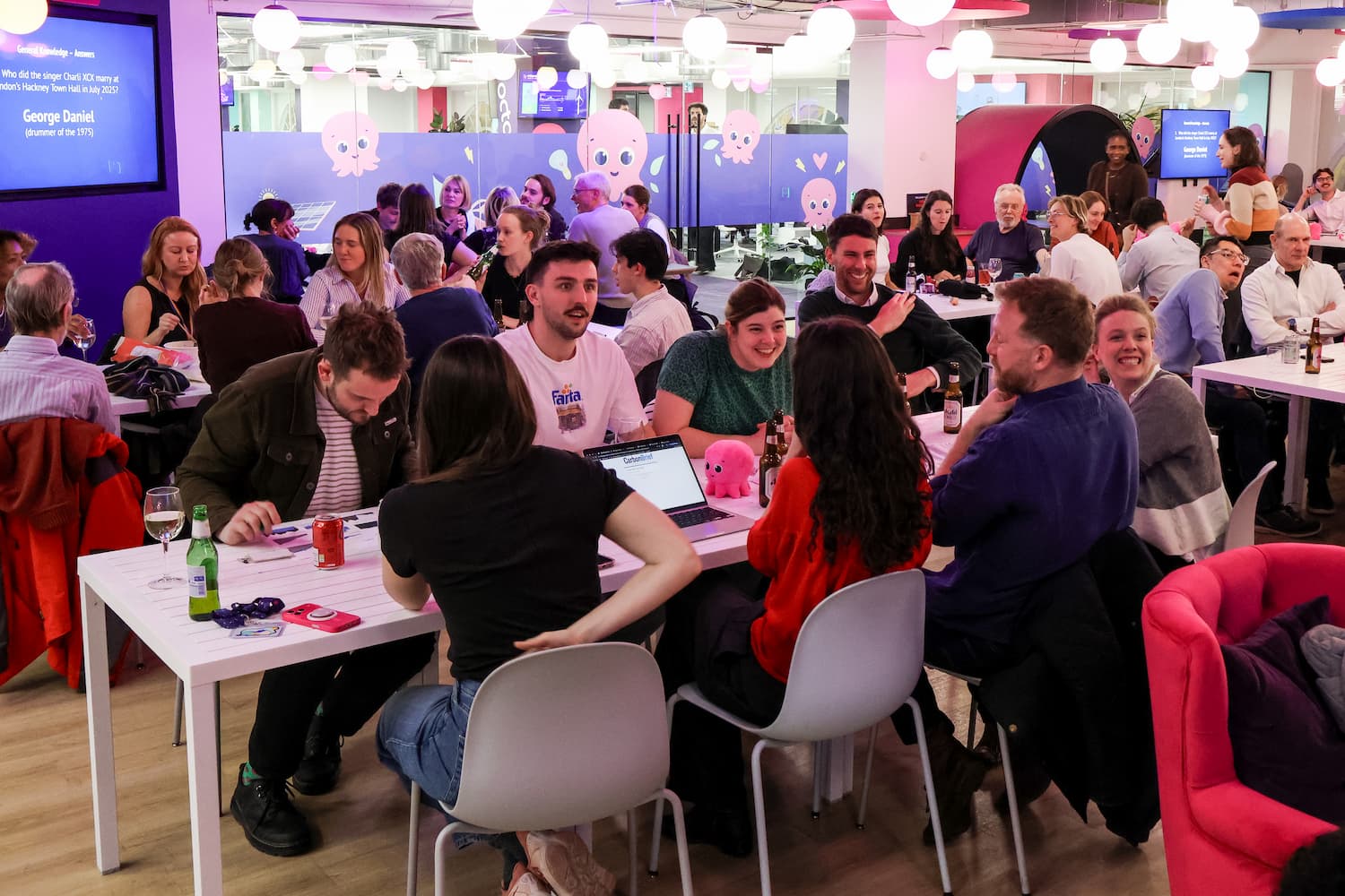 A wide shot of a busy trivia night. Teams are seated at long white tables with laptops and drinks. A screen on the left shows a trivia answer: 'George Daniel' The room features pink and purple decor.