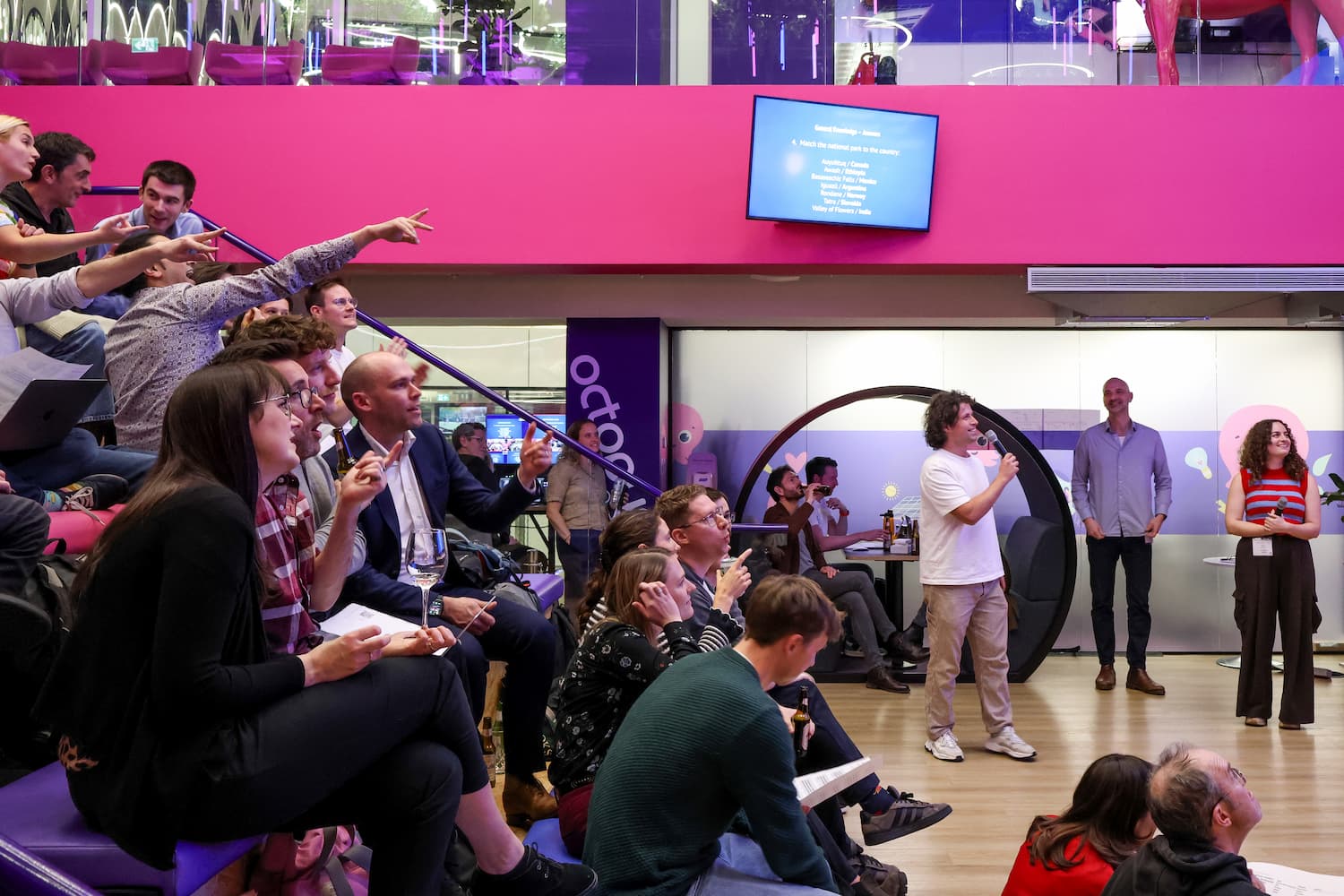 A side view of people sitting on tiered, colorful bleacher seating. Many are pointing toward a screen or a presenter. CB journalist Josh Gabatis stands in the center holding a microphone, leading the event.