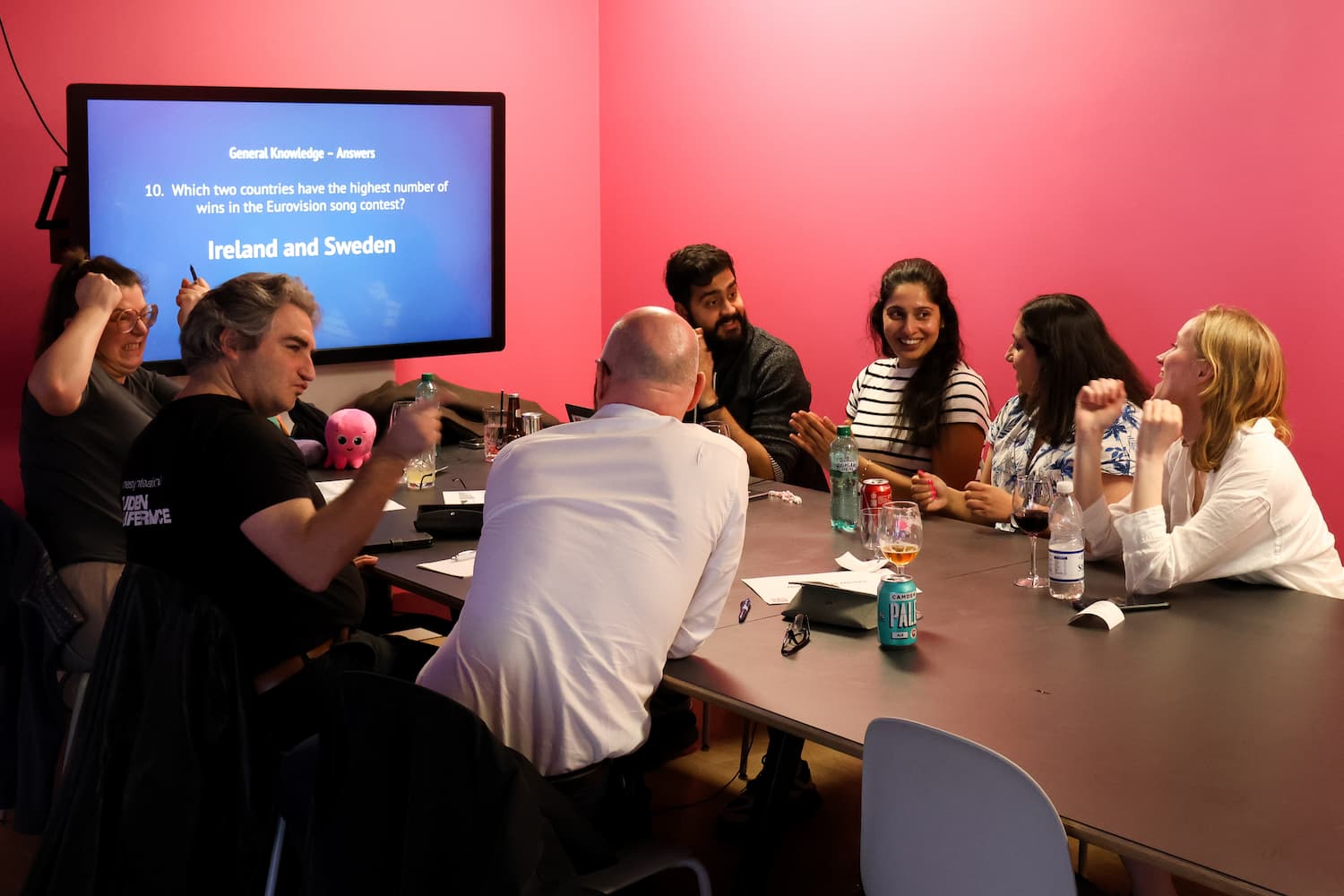 A team sits around a dark table in a room with a bright pink wall. They are cheering and looking at a screen that displays the trivia answer: 'Ireland and Sweden.'