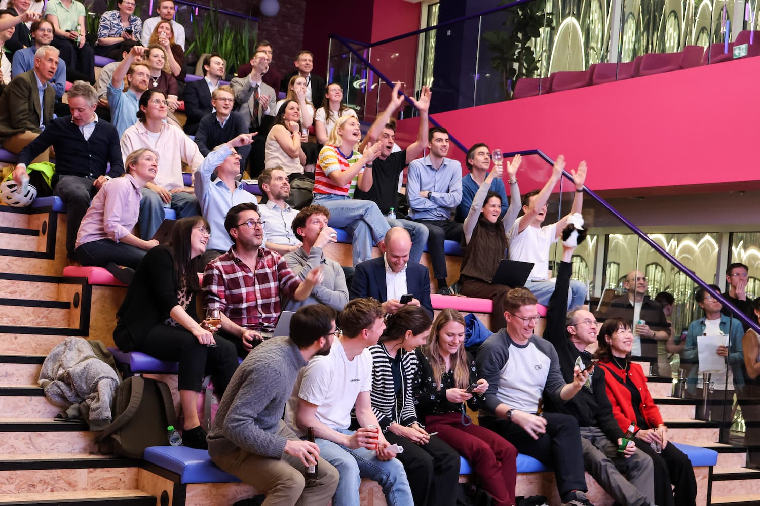 A group of people in a lounge area with bright pink and blue circular ceiling panels. Some are sitting on purple tufted ottomans looking at a laptop, while others stand nearby watching a screen.