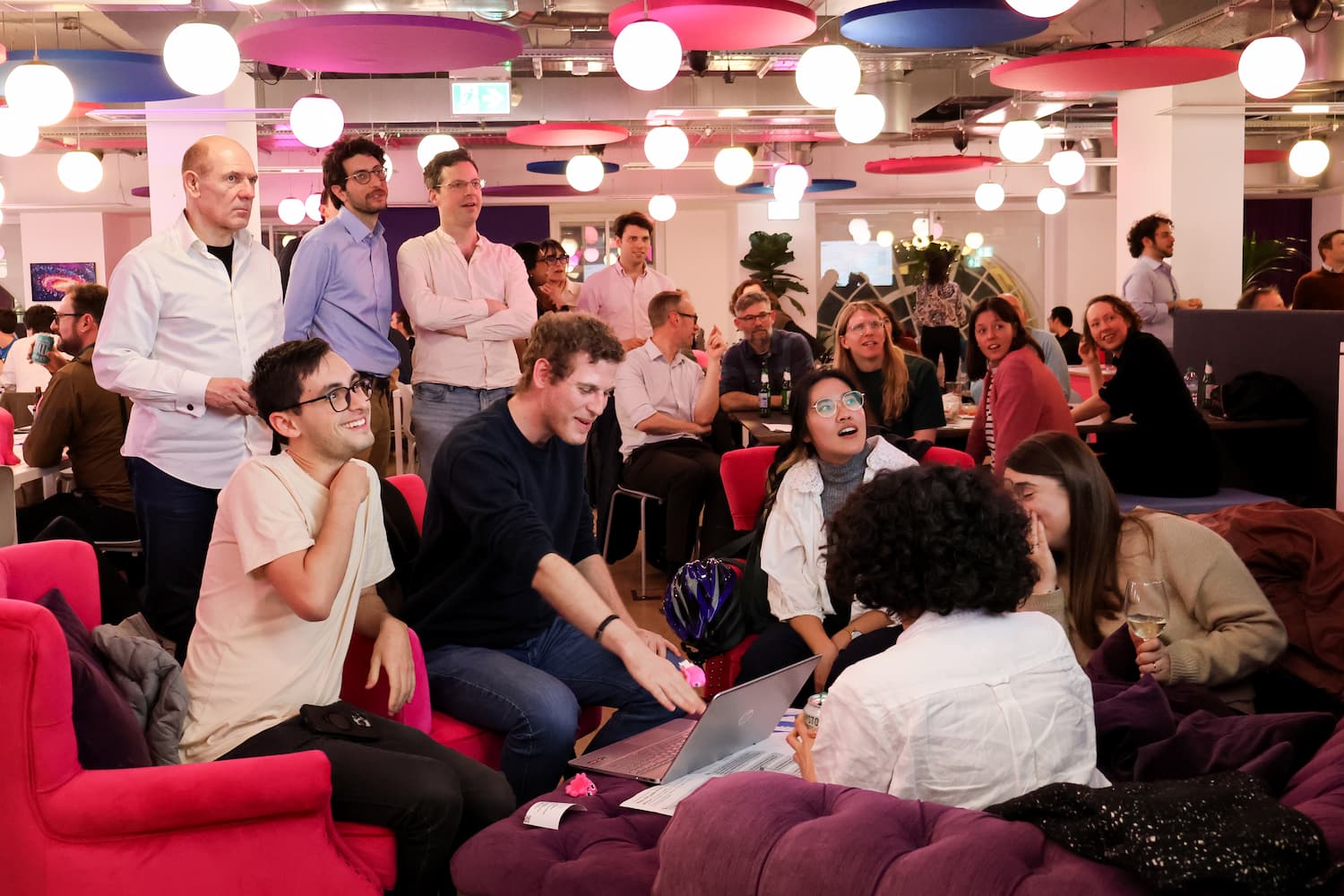 A candid shot of several groups in a modern office lounge. In the foreground, a group sits on a purple tufted sofa looking at a laptop, while others stand behind them watching. The ceiling is decorated with pink, blue, and white circular lights.