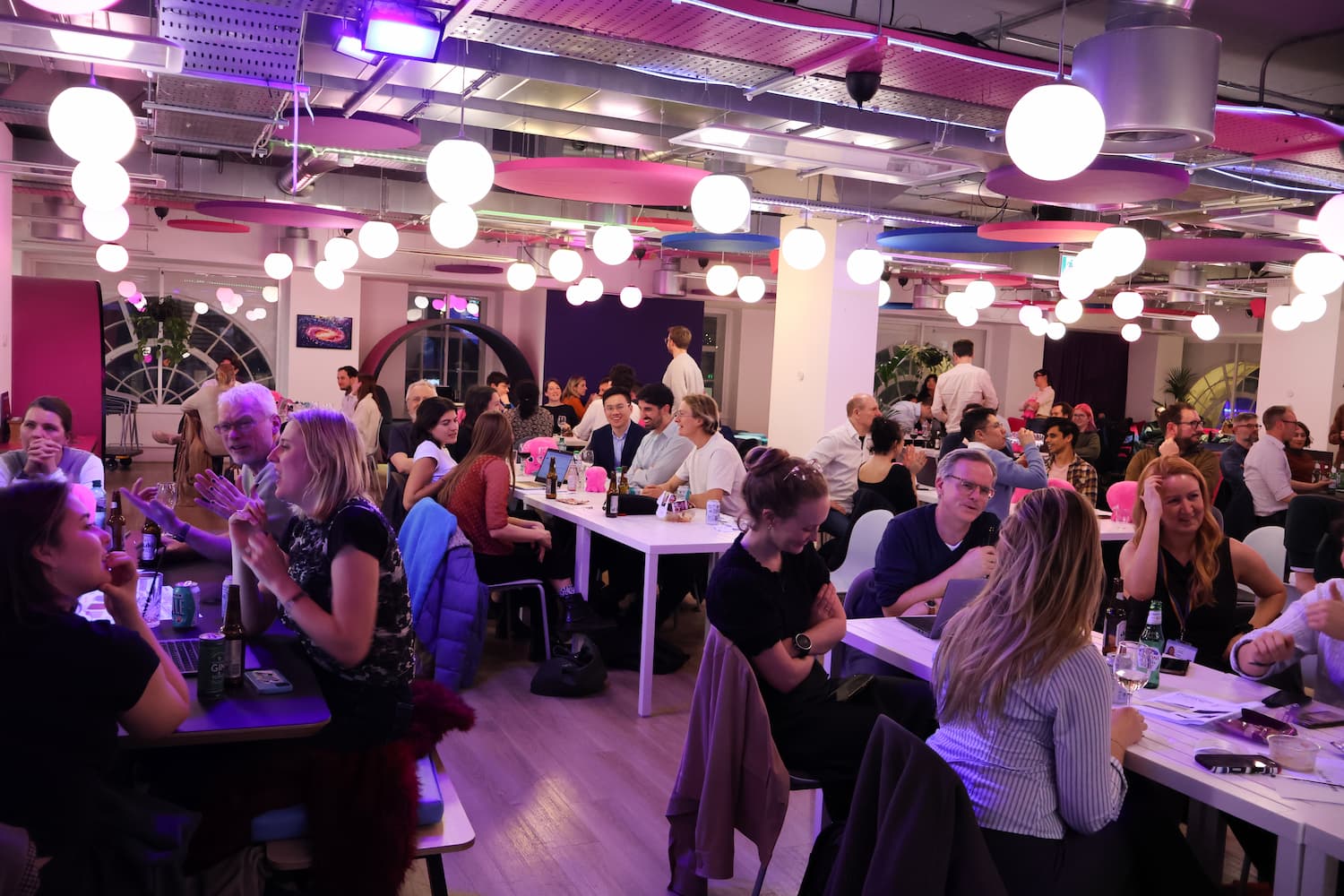 A wide perspective of the Octopus Energy office during the event. Multiple teams are seated at long white tables and smaller dark tables, engaged in conversation and the quiz. The space is filled with warm, glowing globe lights and pink ceiling panels.