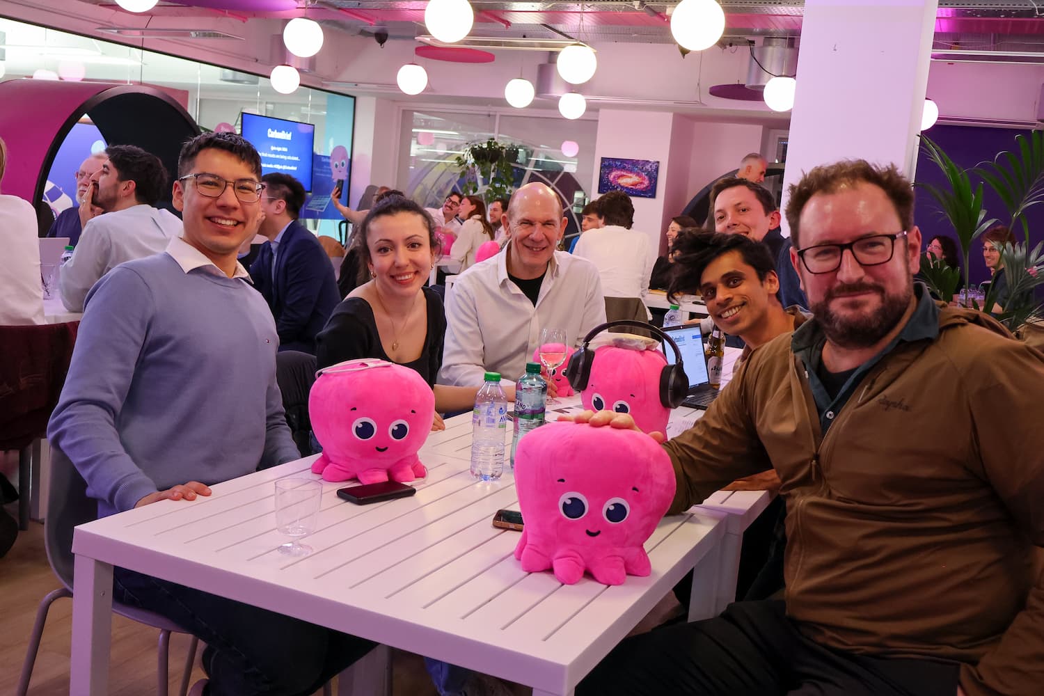 Five people sit at a white outdoor-style table inside the office, smiling directly at the camera. Three bright pink plush octopuses are placed prominently on the table in front of them, one of which is wearing large black headphones.