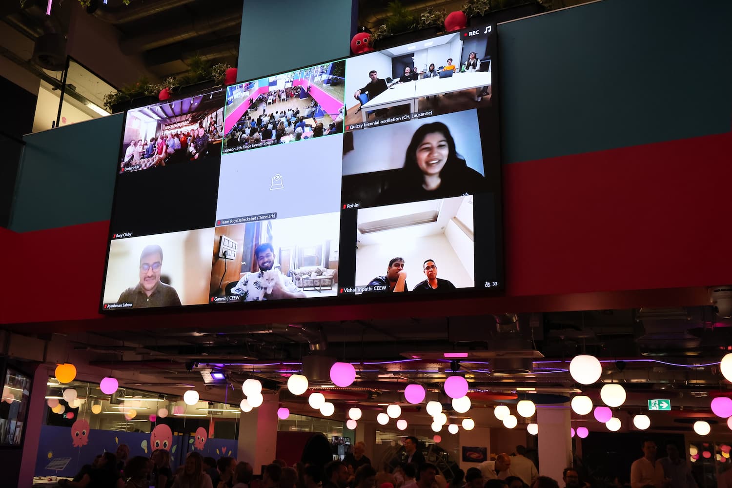 A large digital screen mounted on a red and teal wall displays a video call with several different participants and remote teams. Below the screen, the main event space is visible, filled with people and a dense canopy of hanging globe lights.